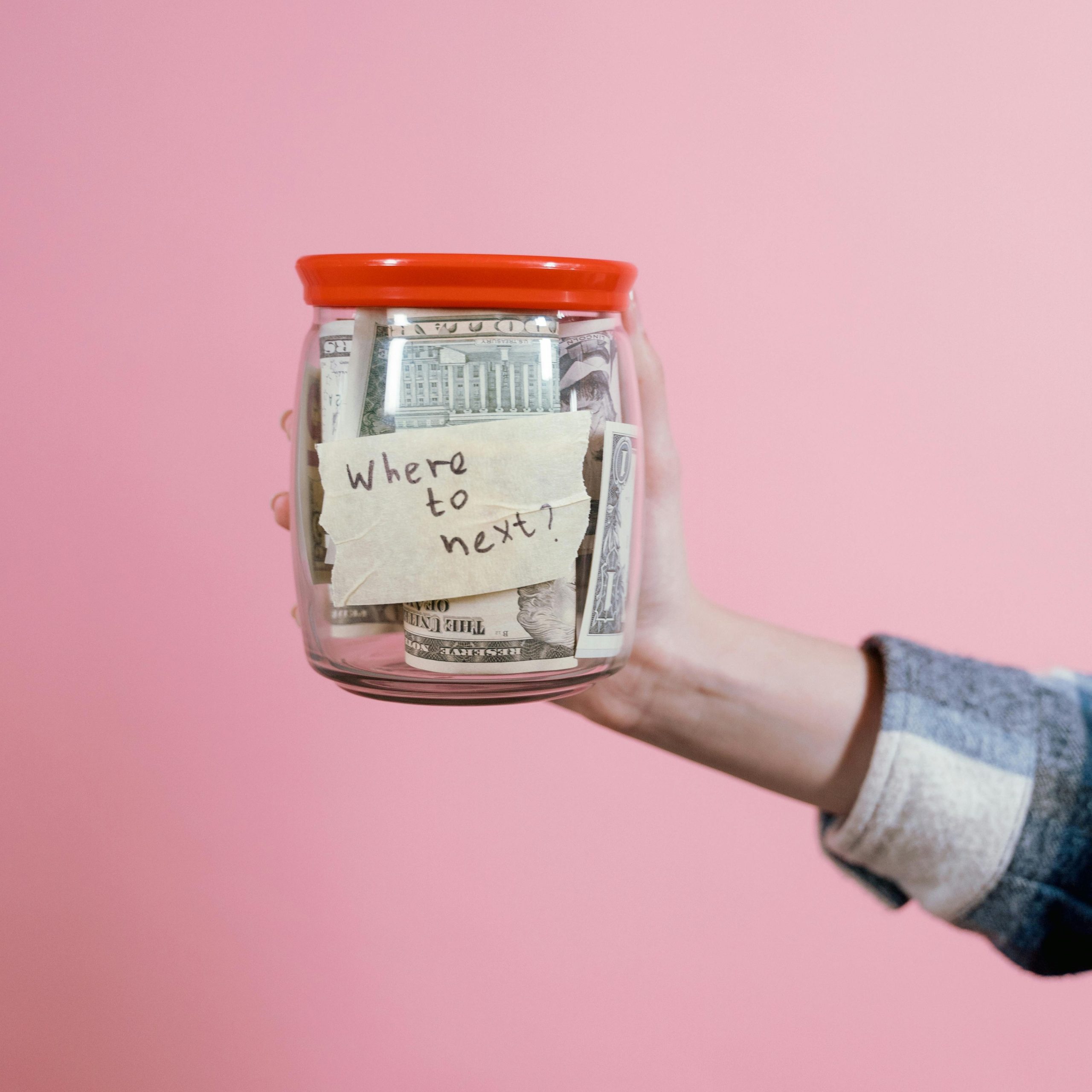 Close-up of a hand holding a savings jar filled with money against a pink backdrop.